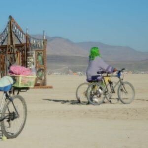Playa Will Provide illustrated by Woman with green hair pushing 2 bikes on playa at Burning Man 2014